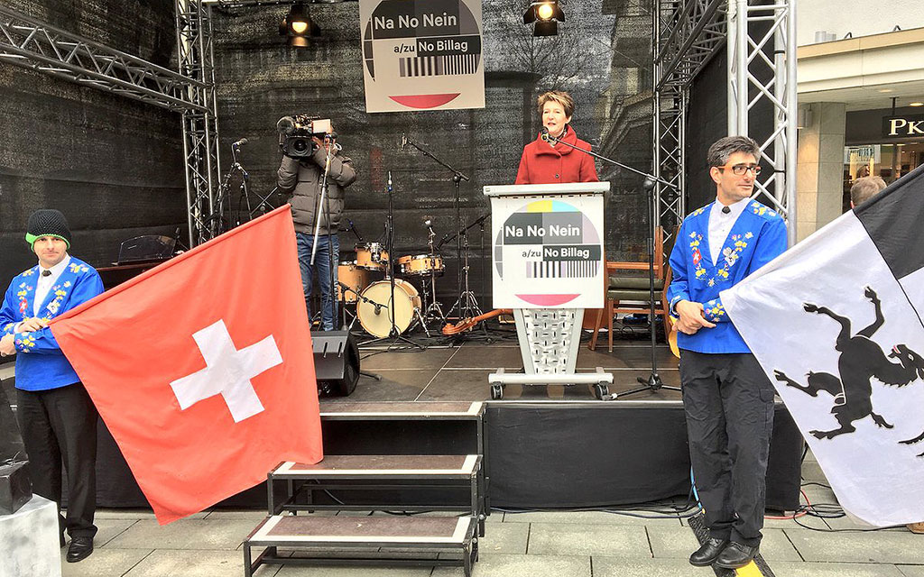 La conseillère fédérale Simonetta Sommaruga prononce son discours à un pupitre placé sur une estrade. De part et d’autre, deux lanceurs de drapeaux.